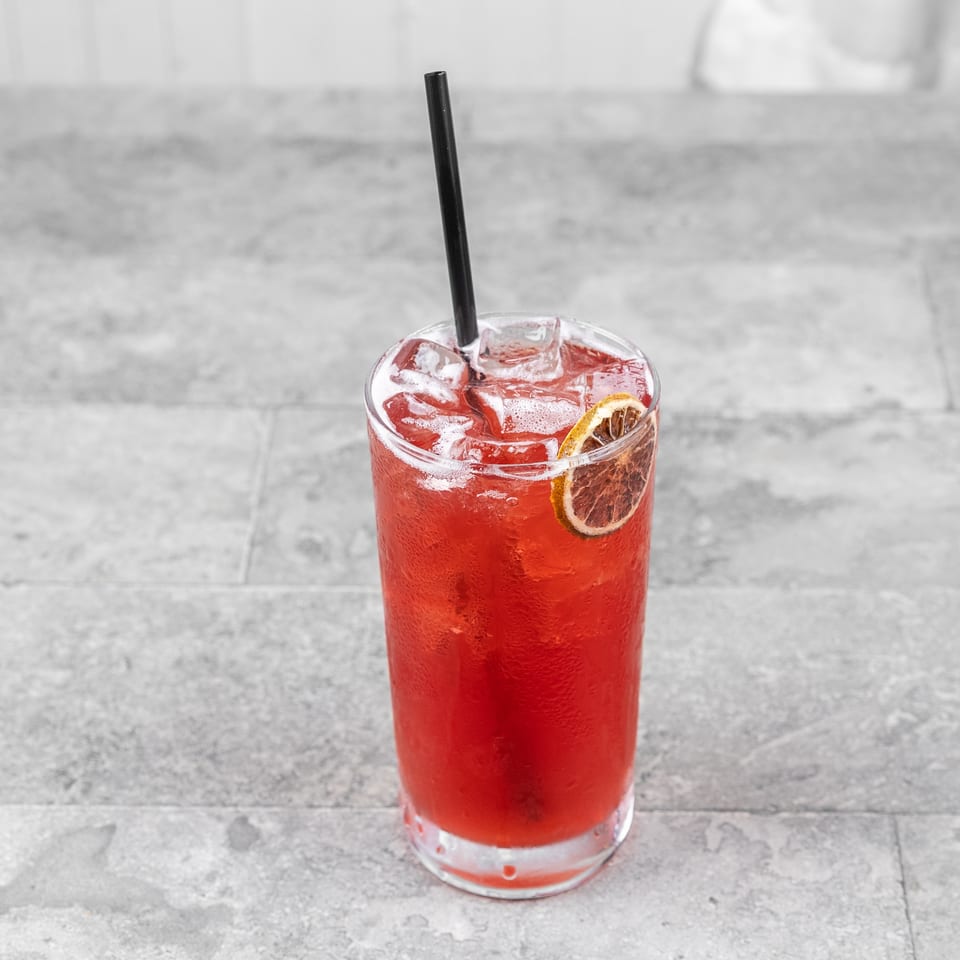 Red iced beverage with a black straw and dried citrus garnish in a clear glass on a gray tabletop.