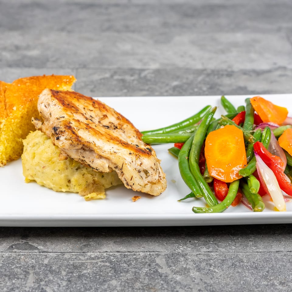 Grilled chicken breast with mashed potatoes, cornbread, and colorful mixed vegetables on a white plate.