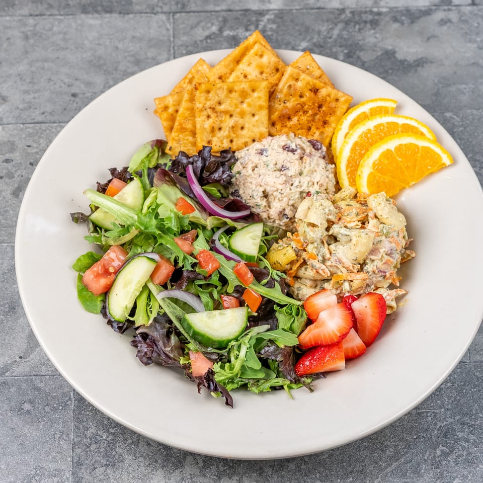 Plate of mixed green salad, creamy deli salads, seasoned crackers, orange slices, and strawberries on a gray tabletop.