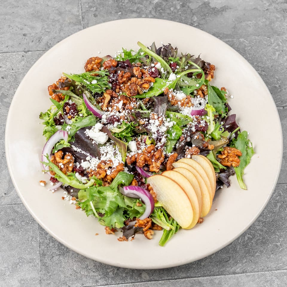 Mixed green salad topped with candied walnuts, dried cranberries, feta cheese, red onion, and sliced apples on a plate.