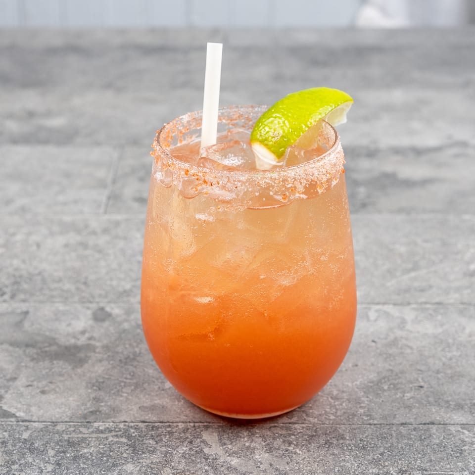 Orange iced cocktail with a chili-salt rim, lime wedge, and straw on a weathered wooden table.