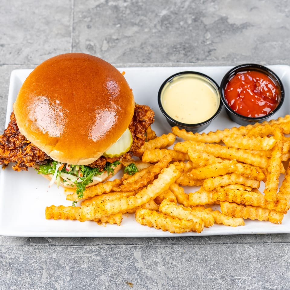 Crispy fried chicken sandwich with pickles and slaw, crinkle-cut fries, cheese sauce, and ketchup on a white plate.