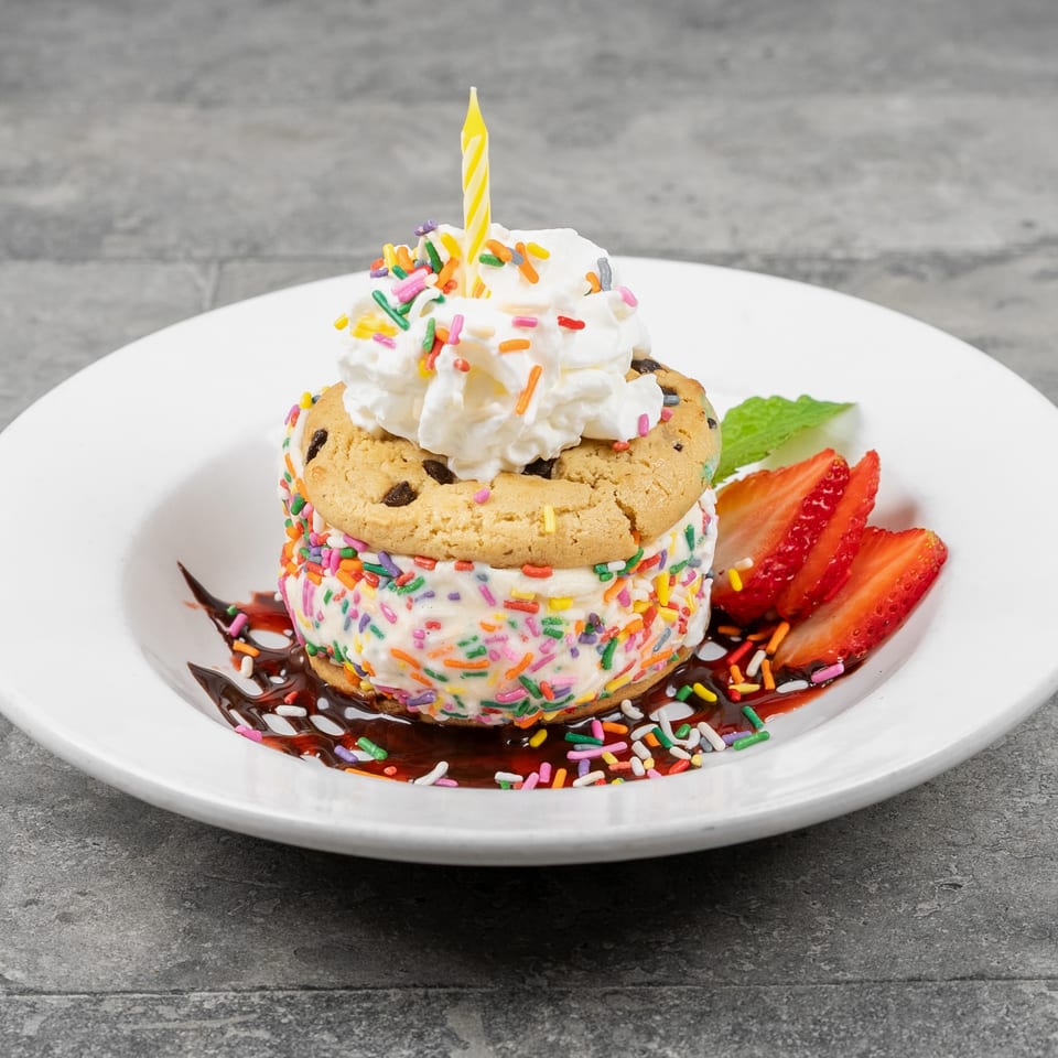 Celebratory ice cream sandwich with chocolate-chip cookies, whipped cream, colorful sprinkles, candle, chocolate sauce, and strawberries.