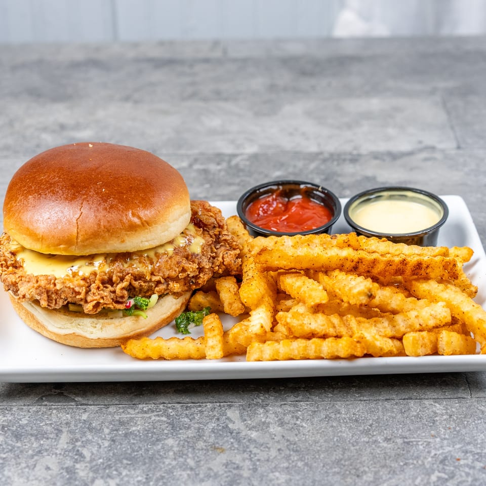 Crispy fried chicken sandwich with crinkle-cut fries, ketchup, and creamy dipping sauce on a white plate.