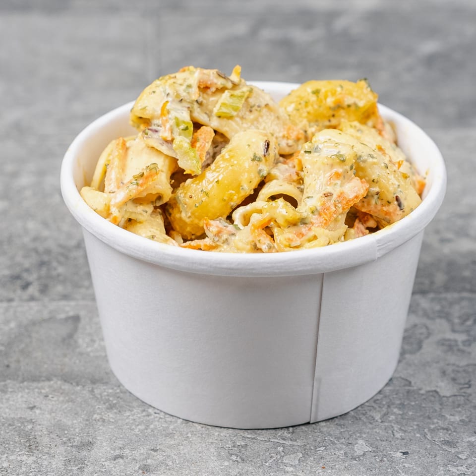 Creamy pasta salad with vegetables and herbs served in a white paper bowl on a gray stone surface.