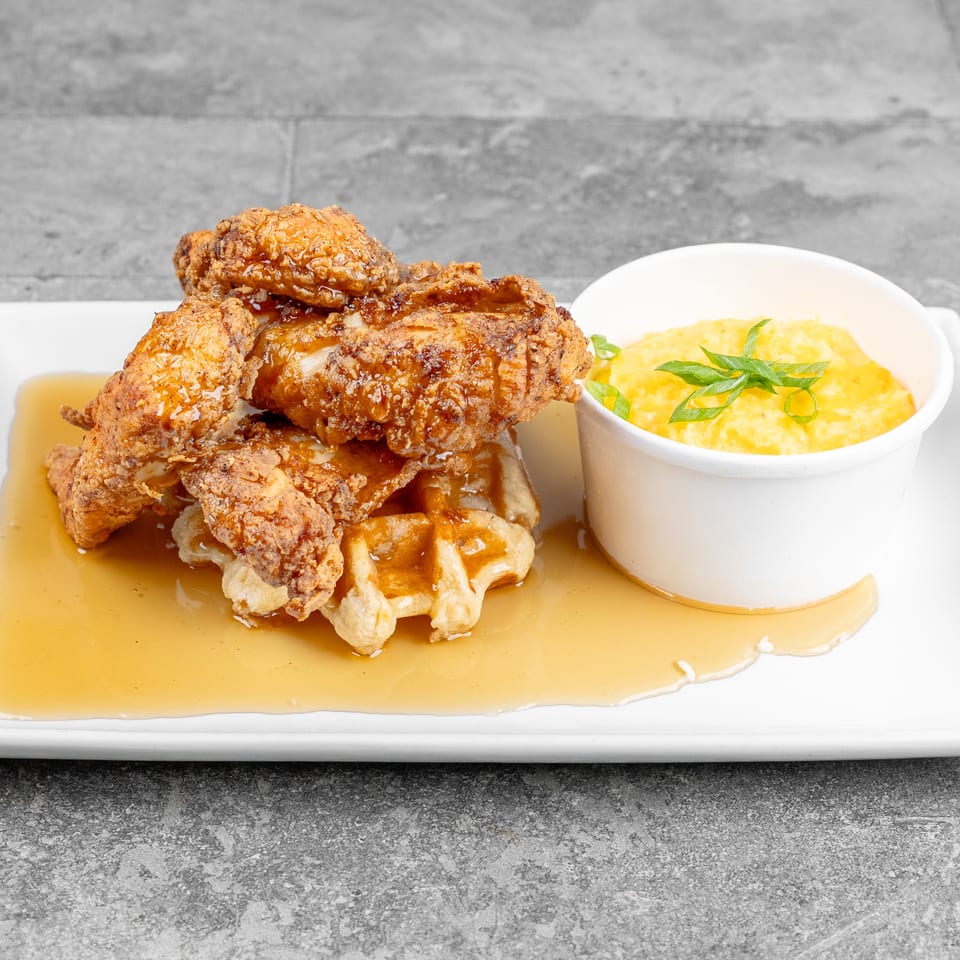 Crispy fried chicken drizzled with syrup sits on a waffle beside creamy yellow grits, garnished with green onions.