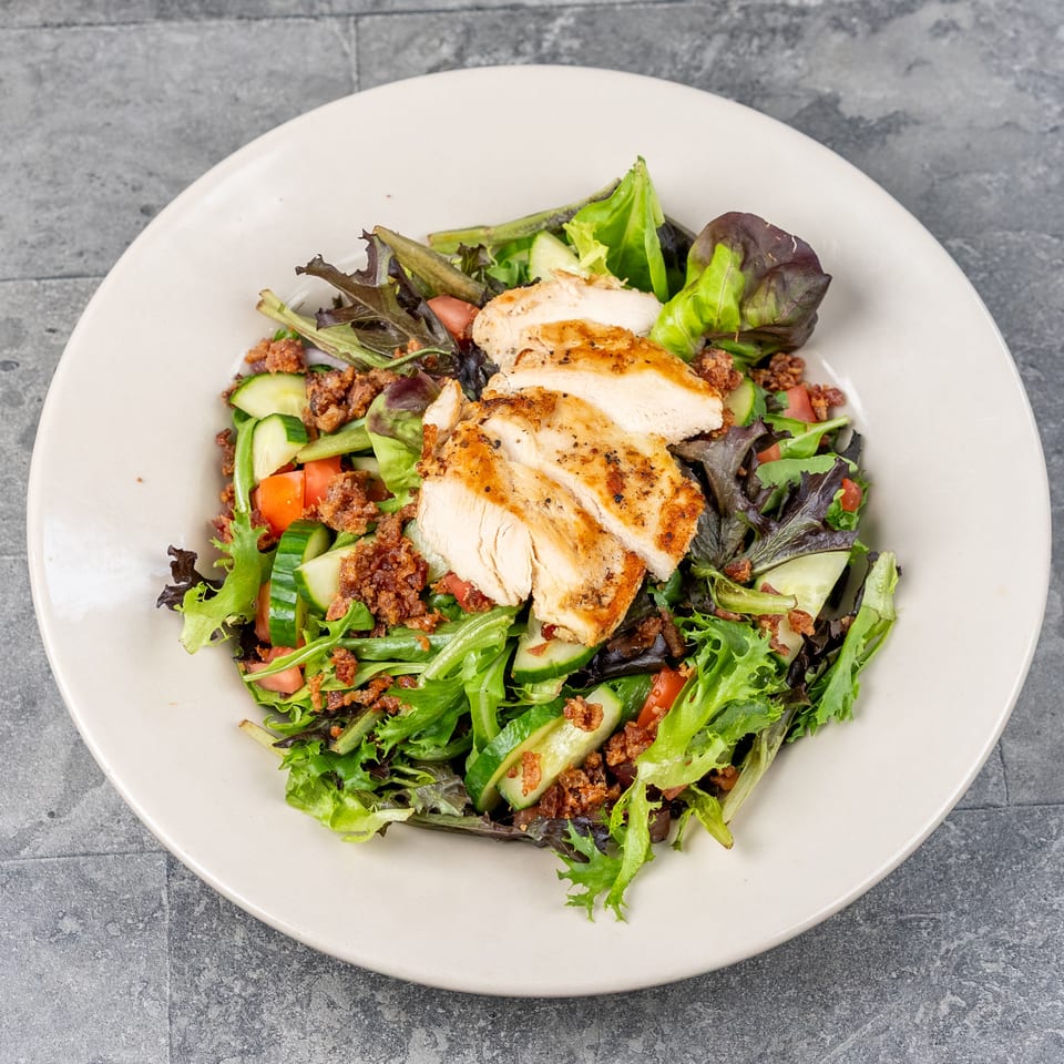 Grilled chicken slices top a fresh mixed-green salad with cucumbers, tomatoes, and crispy bacon on a white plate.