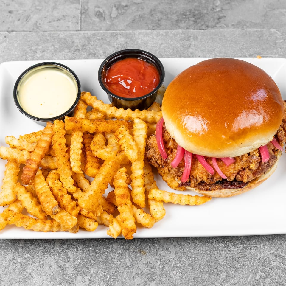 Crispy fried chicken sandwich with pink pickled onions, seasoned crinkle-cut fries, and dipping sauces on a white plate.