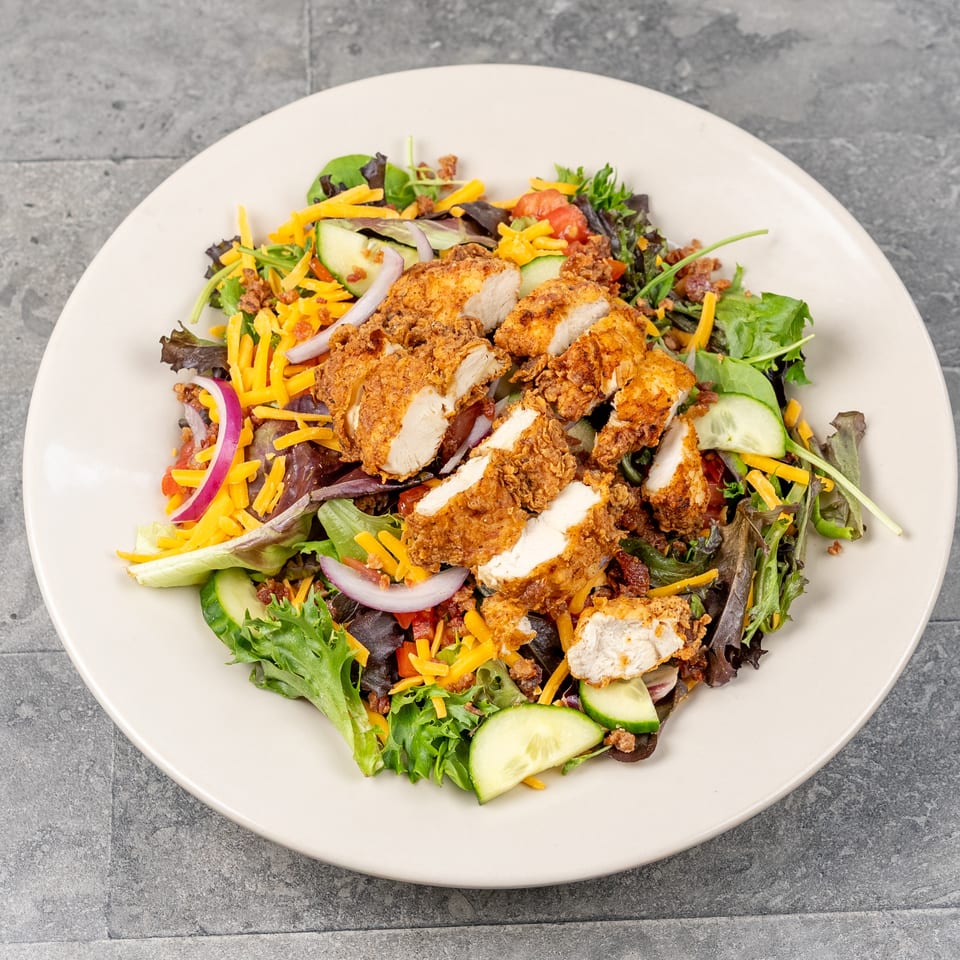 Crispy fried chicken strips top a mixed green salad with cheddar cheese, cucumbers, tomatoes, onions, and bacon.