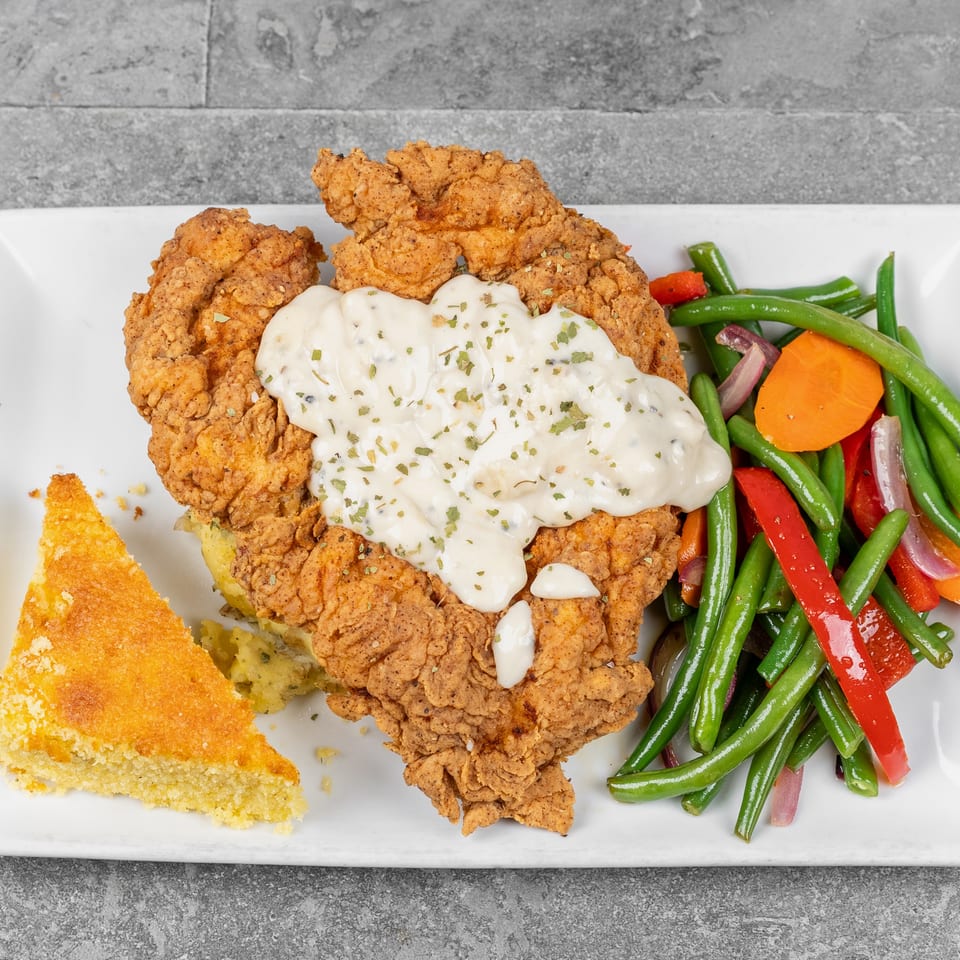 Plate of crispy fried chicken topped with creamy gravy, served with green beans, vegetables, mashed potatoes, and cornbread.