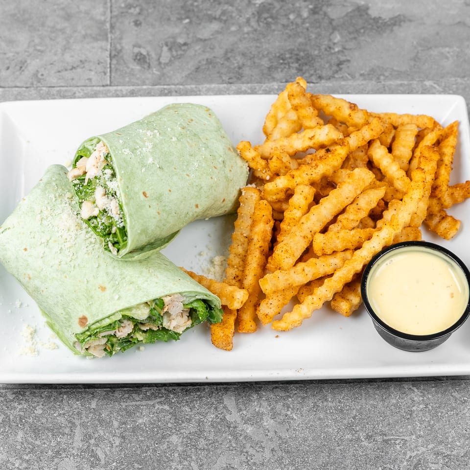 Green chicken salad wrap with leafy greens and cheese served alongside seasoned crinkle-cut fries and dipping sauce.