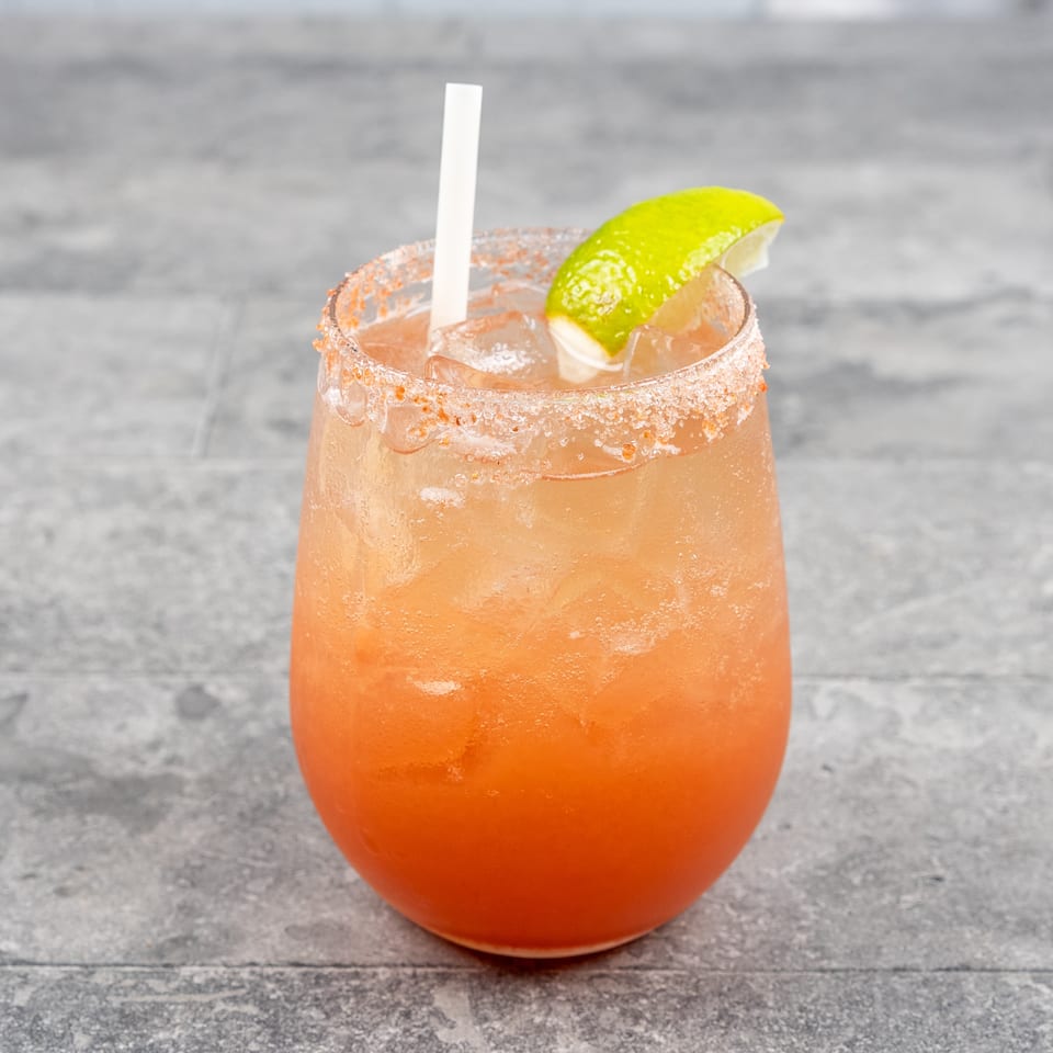 Orange cocktail with ice, a chili-salt rim, white straw, and lime wedge in a short glass on a gray tabletop.